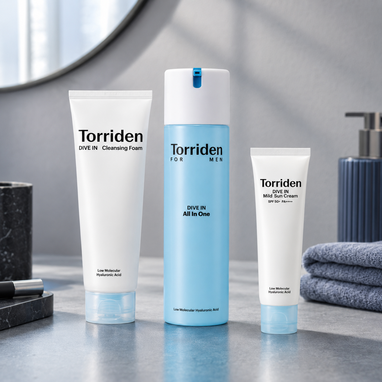 Three Torriden Dive In men's skincare products arranged on a bathroom counter: a white Cleansing Foam tube, a light blue All In One pump bottle, and a white Mini Sun Cream tube, with a folded towel and soap dispenser in the background.