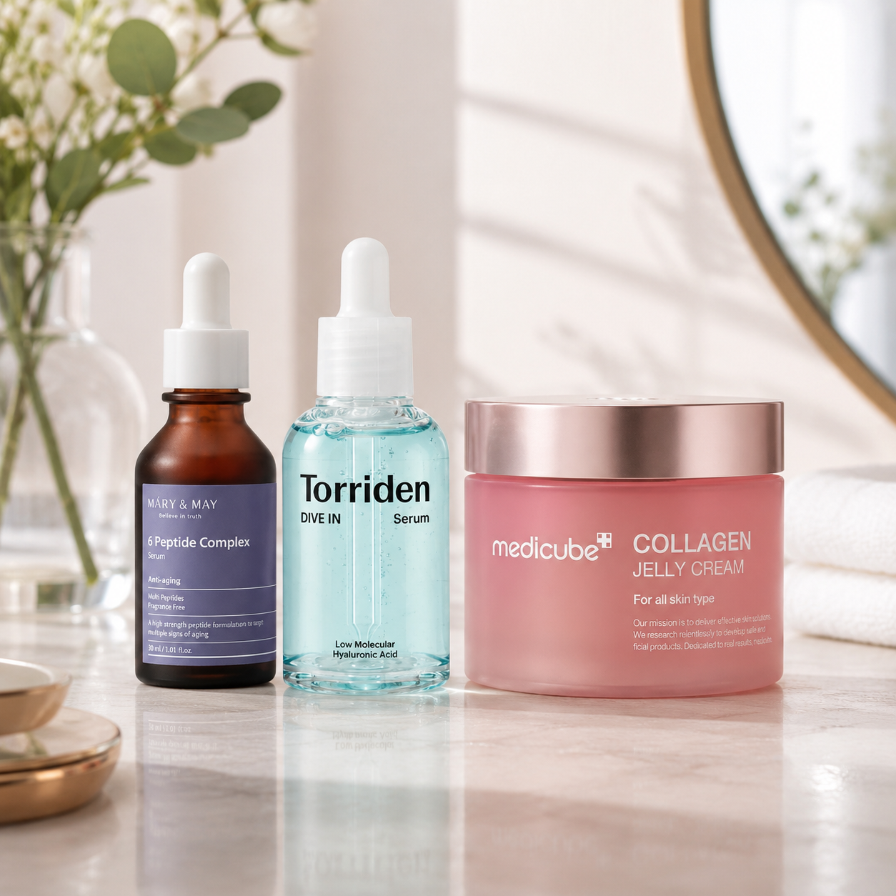 Three skincare products arranged on a marble surface: an amber dropper bottle labeled Nature & May Peptide Complex, a clear dropper bottle labeled Torriden Dive In Serum, and a pink jar labeled medicube Collagen Jelly Cream with a rose-gold lid.